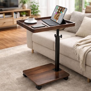 May include: A dark brown, rolling laptop desk with a rectangular tabletop. The desk has a black adjustable stand and a base with wheels. A tablet with a keyboard, a smartphone, and a cup of coffee sit on the tabletop. The desk is next to a beige sofa.