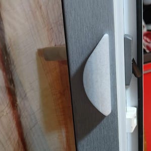 May include: Close-up of a window frame with a gray textured finish. A white, curved handle is attached to the frame. The window's interior reveals a wooden surface.