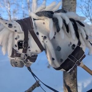 May include: A handmade hobby horse with a white body and black spots. The horse has a brown leather bridle and reins. The mane and forelock are a mix of white, black, and gray yarn. The horse is set against a snowy background.