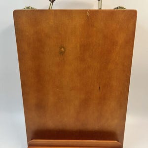 May include: A wooden artist's easel with a brown leather handle and brass fittings. The easel is made of light-coloured wood and has a small shelf at the bottom. It is designed to hold a canvas or artwork.