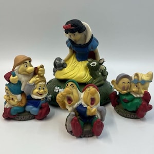 May include: A collection of Snow White and the Seven Dwarfs figurines. The central piece features Snow White with a rabbit and deer. Three smaller groupings of dwarfs are positioned around her, with labels including Grumpy, Happy, and Dopey.