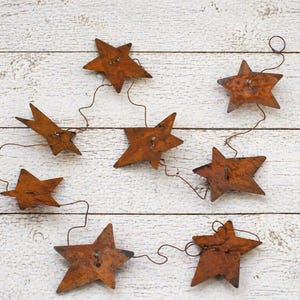 May include: A rustic star garland featuring seven rusty metal stars connected by a thin wire. Each star has a button in the center. The stars are arranged against a white wooden background, creating a charming, country-style aesthetic.