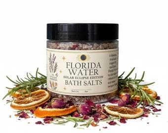 May include: A clear glass jar of Florida Water Solar Eclipse Edition bath salts. The jar is surrounded by dried orange slices, rosemary sprigs, and rose petals. The label features text and botanical illustrations.