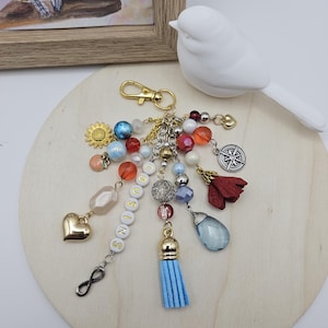 May include: A handmade keychain with a gold clasp and various charms. Charms include a gold heart, a sunflower, beads in multiple colors, a compass, a red flower, and a blue tassel. The word "COUSINS" is spelled out on white letter beads. A white bird figurine is in the background.