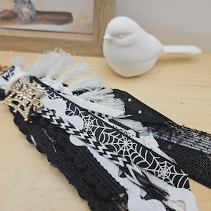 Könnte beinhalten: Ein dekorativer Quasten-Schlüsselanhänger mit Lagen aus schwarzen und weißen Bändern, Spitze und Borten. Das Design umfasst ein schwarzes Band mit weißem Spinnennetz-Muster, eine schwarze Bommelborte, weiße Zackenlitze und einen goldfarbenen Spinnenanhänger mit Strasssteinen.