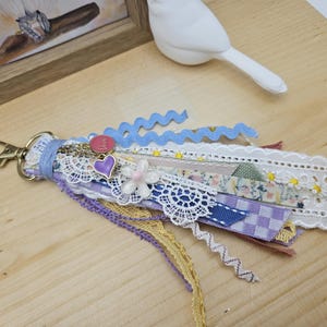 May include: A handmade keychain with a gold-colored clasp. The tassel is made of various ribbons, including lace, floral, and patterned fabrics in shades of blue, purple, and yellow. Embellishments include a heart charm and a white flower.