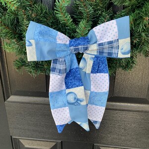 May include: A large, decorative bow made of blue patchwork fabric, featuring various patterns like polka dots, plaid, and floral designs. The bow is attached to a green wreath, creating a festive and handcrafted look. The bow is a light and dark blue color.