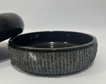 May include: A black stone bowl with a textured, vertically-lined exterior. The interior is smooth and glossy. The bowl is round and sits on a white surface. A portion of a matching lid is visible.