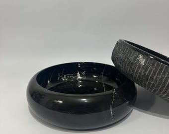 May include: Two black marble bowls. The larger bowl is in the foreground, with a glossy finish and white veining. The smaller bowl is in the background, with a textured, matte finish. Both bowls have a rounded shape.