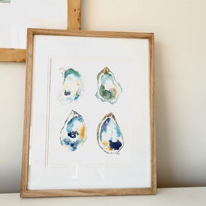 May include: A framed watercolor print featuring four oyster shell illustrations. The shells are painted in shades of blue, green, and gold, with a white background. The artwork is set in a light brown wooden frame.