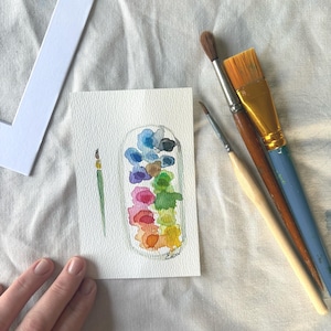 Op de afbeelding: Een aquarel van een pot gevuld met kleurrijke verfspatten. Links van de pot staat een kleine penseel, en rechts drie penselen. De schildering staat op een wit stuk papier, met een witte passe-partout en een witte doek op de achtergrond.