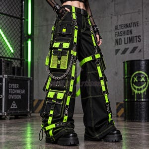black cyberpunk techwear cargo pants with vivid neon green straps, layered utility pockets, mesh panels, metal chains, D-rings, buckles and zipper details. The oversized wide-leg silhouette creates a dramatic cyber goth and industrial punk look, while multiple pocket sections and hanging hardware make these baggy cargo pants ideal for rave, festival, techno and alternative streetwear styling.