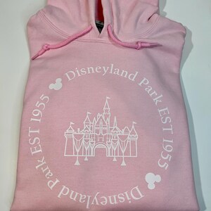 May include: Pale pink hoodie with a drawstring hood. The front displays a white graphic of a castle within a circle, accompanied by the text "Disneyland Park EST 1955". Mickey Mouse head silhouettes are also present.