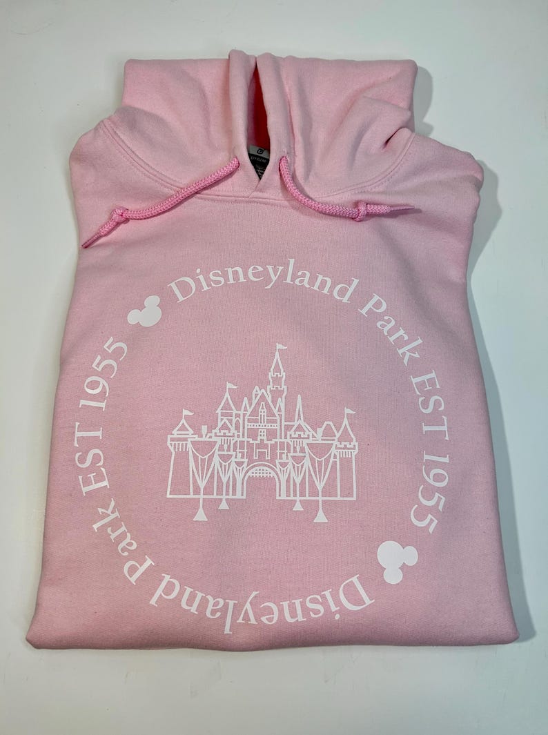 May include: Pale pink hoodie with a drawstring hood. The front displays a white graphic of a castle within a circle, accompanied by the text "Disneyland Park EST 1955". Mickey Mouse head silhouettes are also present.