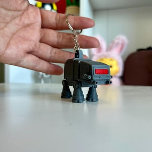 Könnte beinhalten: Ein grauer AT-AT Walker Schlüsselanhänger mit rotem Visier und vier Beinen. Der Schlüsselanhänger ist an einem silbernen Ring befestigt. Der AT-AT ist eine Miniatur-Replik des Fahrzeugs aus dem Star Wars-Franchise.