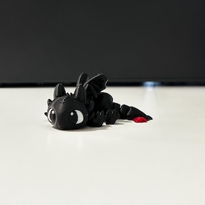 May include: A black dragon figurine with large white eyes and a red accent on its tail. The dragon is lying down, with its wings and legs visible. The figurine is made of a solid material and is resting on a white surface.