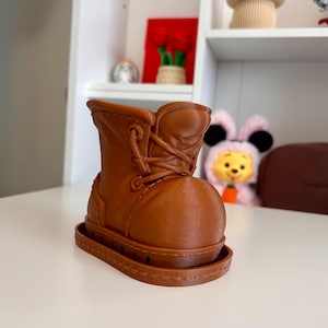 Puede incluir: Una maceta marrón con forma de bota, con una superficie texturizada y un diseño de cordones. La maceta se asienta sobre una base rectangular. La bota es un objeto decorativo.