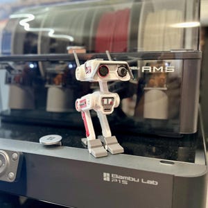 May include: A white and red robot toy with a boxy design, standing on a black surface. The robot has two large eye-like features and antenna. The text "Bambu Lab P1S" is visible on the surface. In the background is a 3D printer.