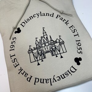 May include: A beige hoodie with a black graphic of a castle and the words "Disneyland Park EST 1955" in a circular design. The hoodie has a drawstring and a front pocket.