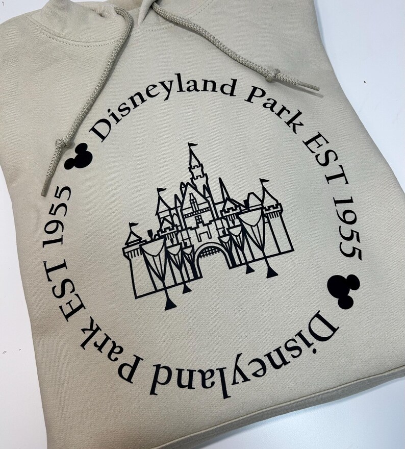 May include: A beige hoodie with a black graphic of a castle and the words "Disneyland Park EST 1955" in a circular design. The hoodie has a drawstring and a front pocket.