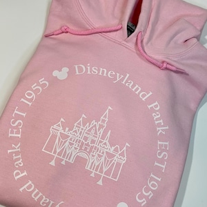 May include: Pale pink hoodie with a drawstring hood. The front displays a white castle graphic and the text "Disneyland Park EST 1955" in a circular arrangement. Includes two Mickey Mouse head icons.