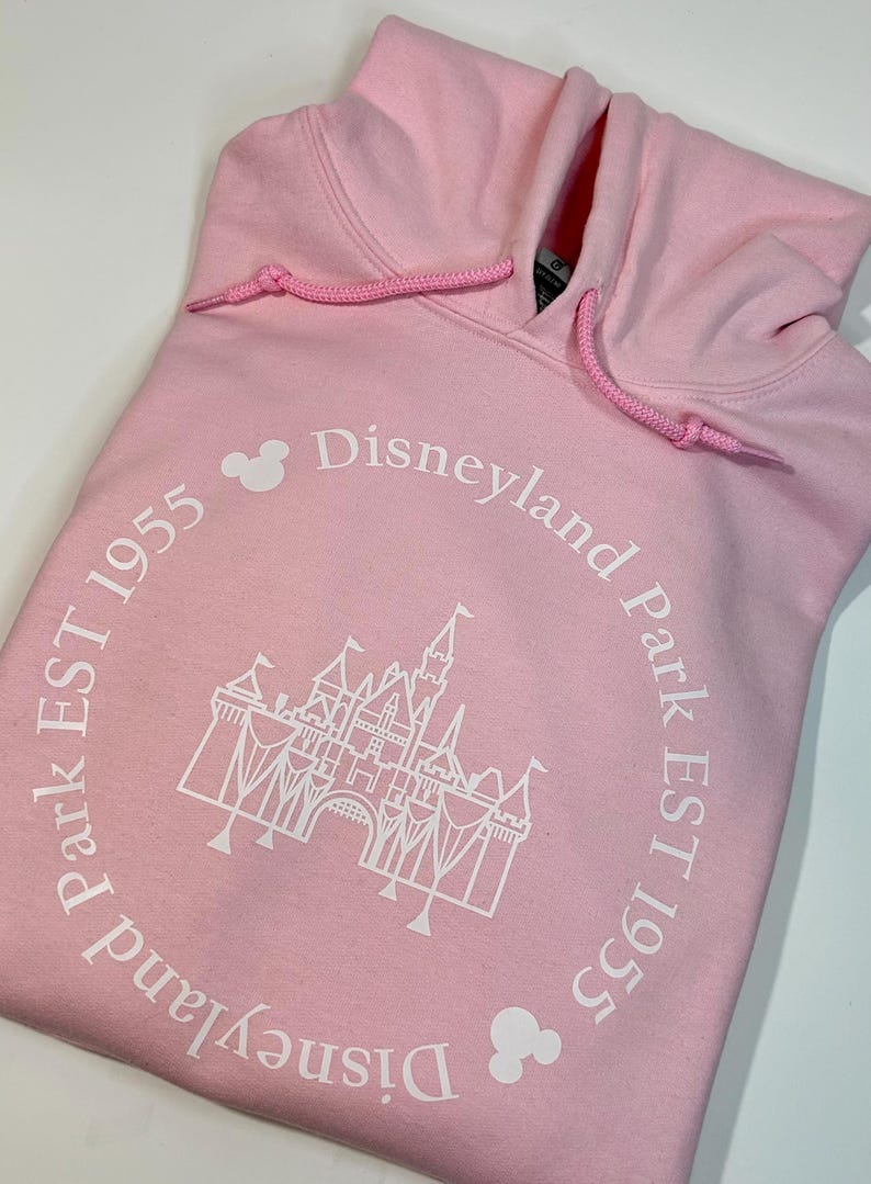 May include: Pale pink hoodie with a drawstring hood. The front displays a white castle graphic and the text "Disneyland Park EST 1955" in a circular arrangement. Includes two Mickey Mouse head icons.