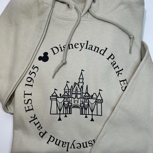 May include: A beige hooded sweatshirt with a drawstring. The front displays a black castle graphic and the text "Disneyland Park Est 1955" in a circular arrangement, with a Mickey Mouse icon. The sweatshirt is folded.