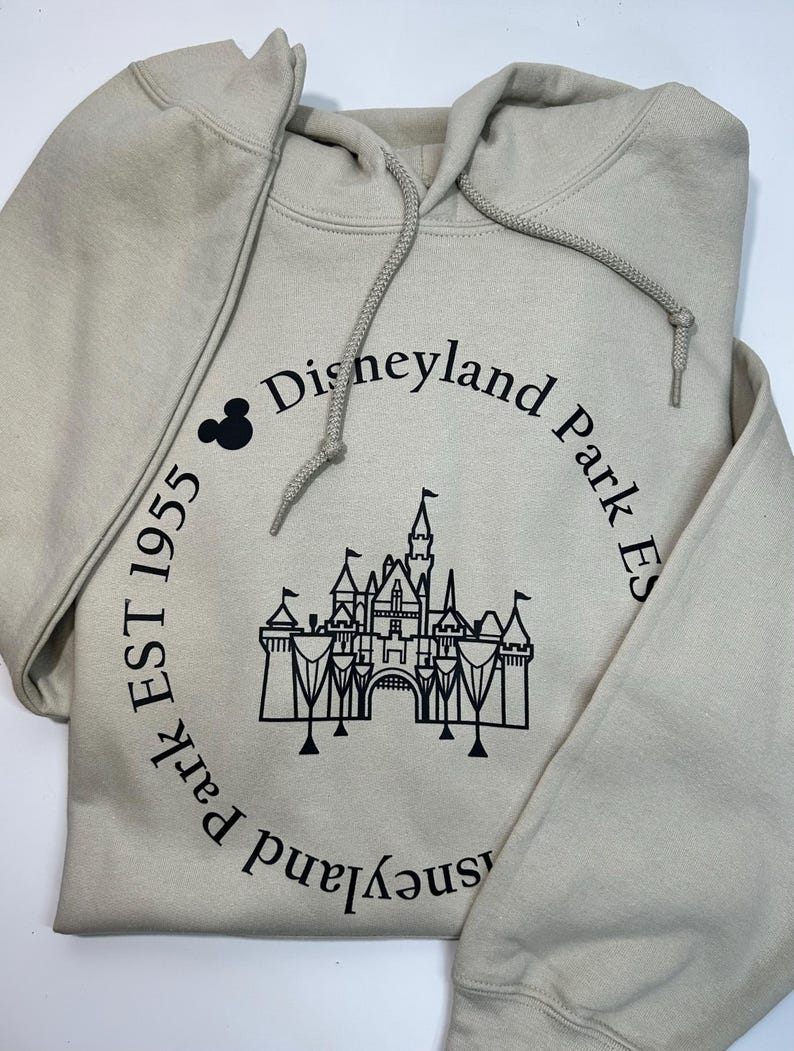 May include: A beige hooded sweatshirt with a drawstring. The front displays a black castle graphic and the text "Disneyland Park Est 1955" in a circular arrangement, with a Mickey Mouse icon. The sweatshirt is folded.