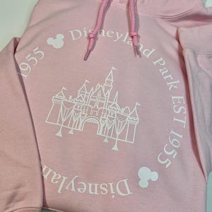 May include: A pale pink hoodie with a drawstring hood. The front displays a white graphic of a castle and the words "Disneyland Park EST 1955" in a circular design. Two Mickey Mouse head icons are also present.