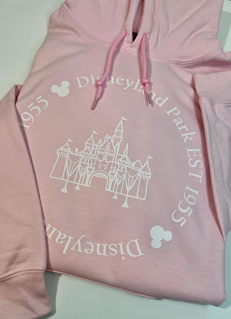May include: A pale pink hoodie with a drawstring hood. The front displays a white graphic of a castle and the words "Disneyland Park EST 1955" in a circular design. Two Mickey Mouse head icons are also present.