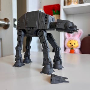 Könnte beinhalten: Ein graues AT-AT-Modell aus Star Wars, das auf einer weißen Oberfläche steht. Das Modell hat detaillierte Beine, einen rechteckigen Körper und ein separates, kleineres Fahrzeugteil. Der Hintergrund umfasst eine Tür, ein Regal mit Gegenständen und ein Stofftier.