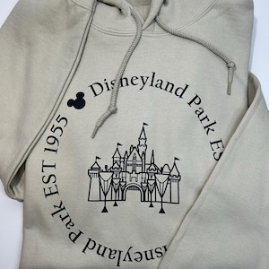 May include: A beige hooded sweatshirt with a drawstring. The front displays a black graphic of a castle and the words "Disneyland Park Est 1955" in a circular design, with a Mickey Mouse icon.