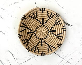 May include: A round woven basket with a spiral center and radiating black lines. The basket is tan and black, with a circular design. The basket is made of natural materials and is likely used for storage or decoration.