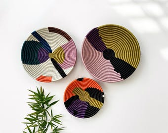 May include: Three woven decorative wall baskets in a variety of sizes. The baskets feature colorful geometric designs in shades of pink, yellow, black, purple, and orange. The baskets are circular and have a spiral pattern.