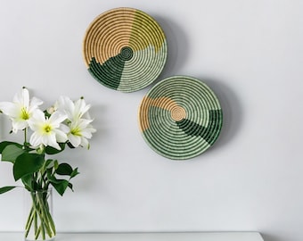 May include: Two round woven wall baskets with spiral designs. Each basket features sections of tan, green, and dark green. The baskets are mounted on a white wall, with a vase of white flowers on a white surface below.
