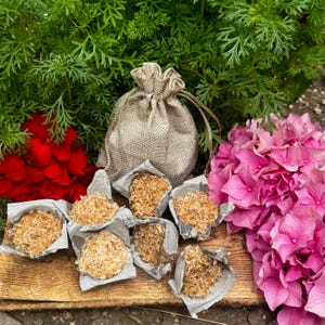 May include: A beige hessian drawstring pouch sits behind six small, silver-lined paper cups filled with shredded light brown wood fibre. The arrangement is placed on a wooden board between a cluster of red flowers and a cluster of pink flowers.