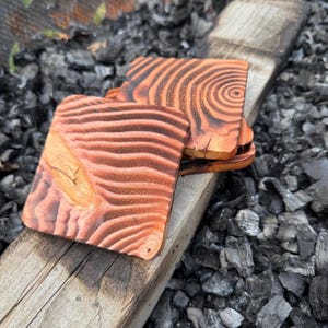 Può includere: Set di quattro sottobicchieri quadrati in legno con un motivo a venature naturali. I sottobicchieri sono di un caldo colore marrone rossiccio con linee di venature marroni più scure, impilati su una tavola di legno.