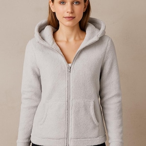 Op de afbeelding: Een lichtgrijze fleece hoodie met ritssluiting, capuchon en twee voorzakken. De jas heeft een ritssluiting over de hele lengte en een zachte, getextureerde uitstraling. Het model draagt er een witte top onder.