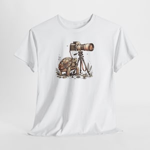 May include: White t-shirt featuring a whimsical illustration of a turtle with a camera on a tripod. The turtle is wearing a backpack and is positioned next to the camera. The camera has a long lens. Ferns and small rocks are in the foreground.