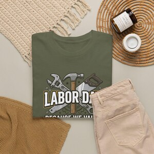 Puede incluir: Una camiseta verde oliva con un estampado gráfico de un martillo, una llave inglesa y una sierra cruzados. El texto dice LABOR DAY BECAUSE WE VALUE. El diseño incluye elementos de engranaje estilizados y cabezas de tornillo en blanco, amarillo y gris.