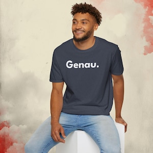 May include: Navy blue t-shirt with the word "Genau" in white block letters. The shirt is made of a soft material and is being worn by a person. The person is wearing blue jeans.