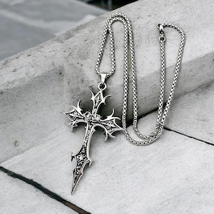 Op de afbeelding: Een zilverkleurige hangerketting met een gotisch kruisontwerp. Het kruis heeft ingewikkelde details en hangt aan een bijpassende ketting. De hanger heeft een puntige onderkant en decoratieve randen.