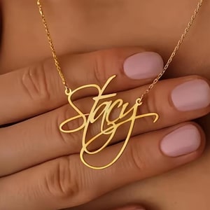 Peut inclure: Un collier doré avec le nom "Stacy" écrit en cursive. Le collier est tenu par une main avec du vernis à ongles rose. La chaîne est fine et le pendentif est en une seule pièce de métal.