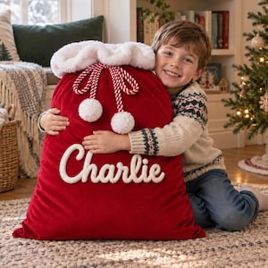 Puede incluir: Un saco de Papá Noel grande de terciopelo rojo con un borde de felpa blanca en la parte superior, dos pompones blancos unidos a cordones trenzados rojos y blancos, y el nombre Charlie bordado en letras cursivas blancas al frente.