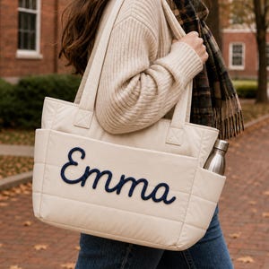 May include: A cream-colored quilted tote bag featuring the name Emma embroidered in dark blue script on the front exterior pocket. The bag includes two top handles and a side pocket containing a metallic silver water bottle.