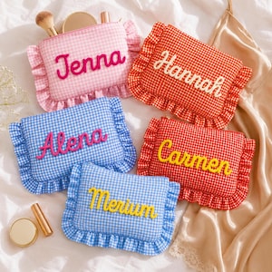 May include: Five rectangular zippered pouches with ruffled edges and gingham patterns in pink, orange, red, and blue. Each pouch features a name embroidered in cursive script: Jenna, Hannah, Alena, Carmen, and Merium, using pink, white, or yellow thread.