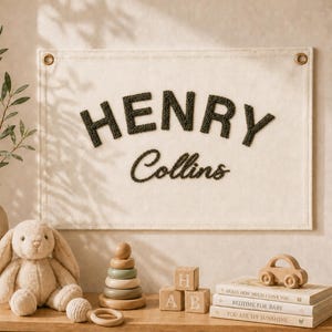May include: A rectangular off-white fabric wall banner featuring the name HENRY COLLINS in dark green textured lettering. The banner has metal grommets in the top corners for hanging and is displayed with nursery decor including a plush rabbit.
