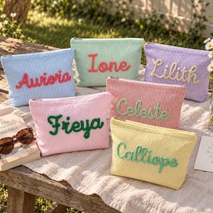 Pode incluir: Seis estojos retangulares com zíper feitos de tecido seersucker listrado em azul claro, verde menta, roxo, rosa, vermelho e amarelo. Cada estojo apresenta um nome bordado em uma cor de fio contrastante: Aurora, Ione, Lilith, Freya, Celeste e Calliope.