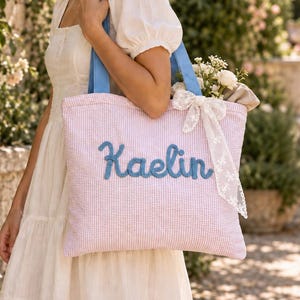 Puede incluir: Un bolso tote de seersucker en color rosa claro y blanco con el nombre Kaelin bordado en letras cursivas azules. El bolso tiene asas azules y está decorado con un lazo de encaje blanco atado en la parte superior derecha.