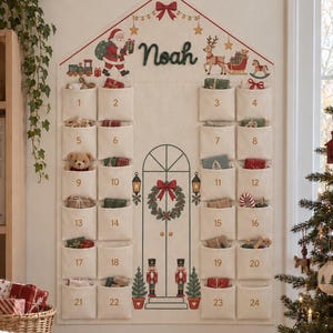 May include: A cream-colored fabric advent calendar hanging on a wall, featuring the name Noah, twenty-four numbered pockets, and illustrations of Santa, a reindeer, a wreath-adorned door, and two nutcrackers with green Christmas trees.